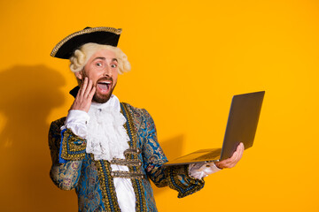Young man in historic costume holding a laptop against a yellow background with a surprised...