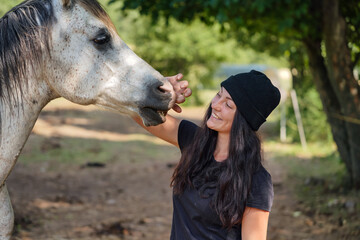 Young woman in casual work clothes standing near her white Arabian horse on ranch farm