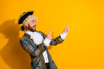 Cheerful young man dressed as an aristocratic nobleman pointing to the side against a bright yellow...