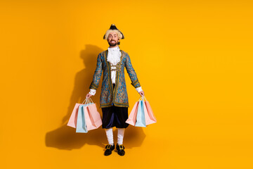 Elegant young man in historic nobleman costume holding shopping bags against vibrant yellow...