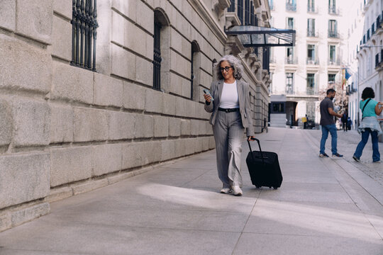 Full length, Elegant senior businesswoman in gray suit walks confidently with her suitcase, multitasking on smartphone.Female embodies professionalism, success, and adaptability in a globalized world