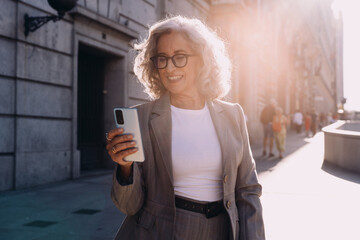 Successful senior businesswoman checks smartphone with smile, enjoying bright city atmosphere....