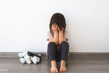 little child 2 year sits on the floor against white wall, covering her face with her hands. plush...