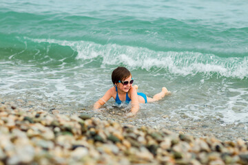 A small, beautiful girl in a blue swimsuit and sunglasses, swimming on the beach
