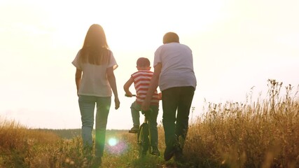 Family teaches little boy to ride bicycle at countryside at sunset. Happy family of mother father and son enjoys activity in wild field. Man helps son to ride bike for first time at family leisure