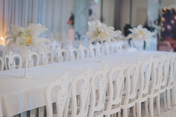 A beautifully arranged banquet hall features white chairs and elegant floral centerpieces on each table. The soft lighting enhances the ambiance, preparing for a special event