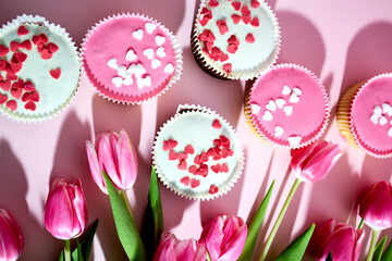 Sponge cupcakes with pink and white icing and heart-shaped sugar sprinkles on a pink background next to tulip flowers