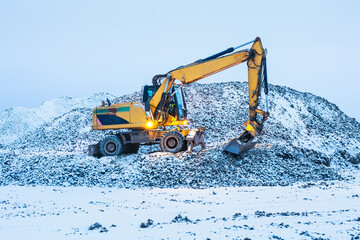 A excavator on a winter morning at the construction site, standing on a pile of dirt covered with snow