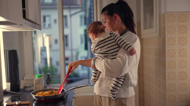 Mother holding baby while cooking in kitchen, stirring food in frying pan, multitasking with child in arms, everyday parenting responsibilities in home setting