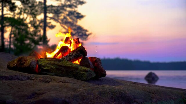 A cozy campfire burns on a rocky lakeshore during the white nights of summer solstice in Finland. Video with nature sounds for relaxation and meditation.