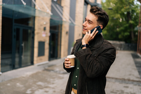 Handsome bearded young man drinking tasty coffee and talking on smartphone while walking in city street by modern urban buildings, enjoying pleasant conversation during break outdoors alone.