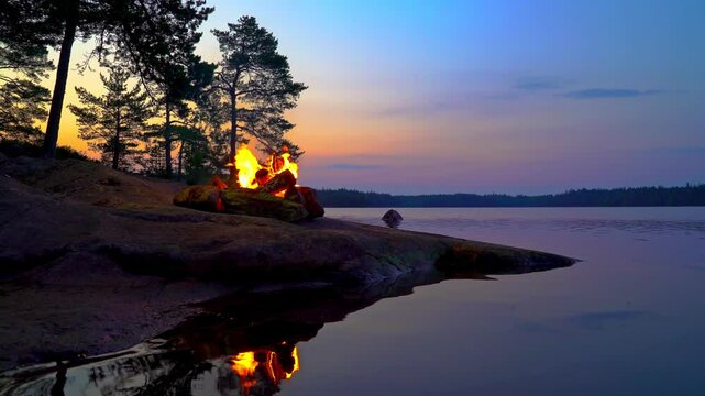 A cozy campfire burns on a rocky lakeshore during the white nights of summer solstice in Finland. Video with nature sounds for relaxation and meditation.