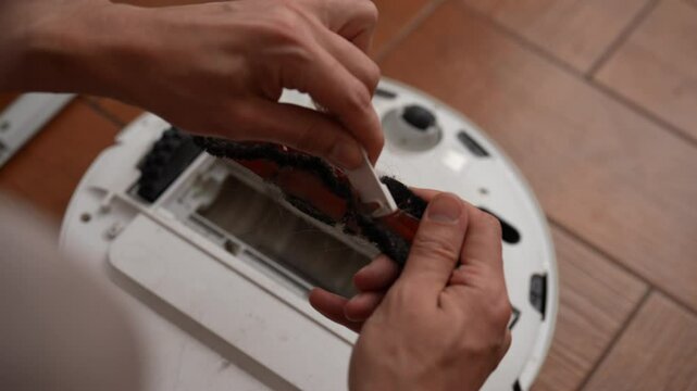 Top view of maintenance process for robotic vacuum cleaner, male carefully removing entangled hair strands and accumulated debris from rotating brush using small knife, close-up, slow motion.