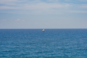 Fototapeta premium Vast Mediterranean Sea with a solitary sailboat on the horizon under light clouds on a clear day.