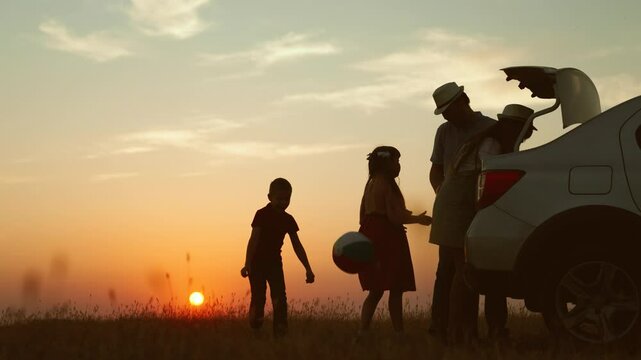 Playful family at sunset by car. cheerful children parents going on travel road trip by automobile packing baggage putting luggage in trunk kids playing ball. Packing preparation for family picnic.