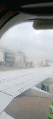 A foggy morning view from an airplane window, showing the aircraft wing and airport terminal. Ground crew prepares for departure as mist creates a dreamy, surreal atmosphere, enhancing the travel expe