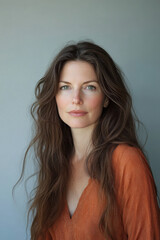 Naklejka premium Portrait of a Caucasian woman with long wavy brown hair, wearing an orange top, against a soft gray backdrop.