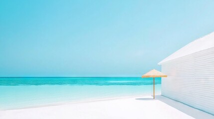Fototapeta premium An umbrella sits on the beach adjacent to a white building and a blue ocean behind, with a blue sky above