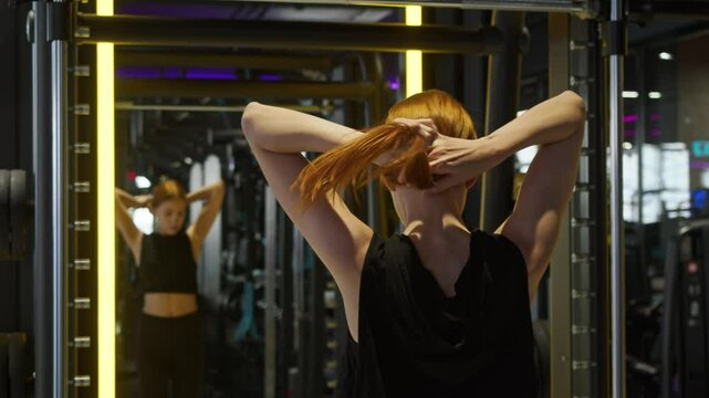 Woman with long, luxurious red hair gathers her curls into pigtail secured with an elastic band before starting an intense workout in gym.