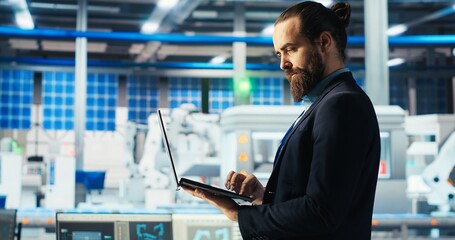 Fototapeta premium Man in photovoltaics factory monitoring system performance on laptop. Solar panels manufacturing plant employee using notebook to track energy production and efficiency, camera B close up