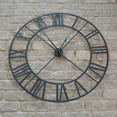 A large metal wire frame clock with roman numerals hangs on a grey stone wall.