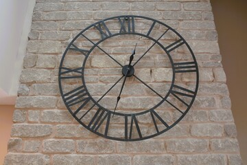 A large metal wire frame clock with roman numerals hangs on a grey stone wall.