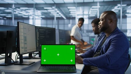 Employees using green screen laptop, working in server farm to optimize it for data processing requirements. Close up of mockup notebook on desk next to workers coding in data center, camera A