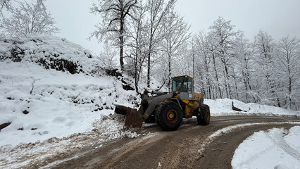 A heavy-duty loader clears snow from a winding mountain road, surrounded by frost-covered trees under an overcast sky