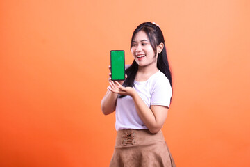 Naklejka premium A smiling woman is presenting a smart phone gadget with a green screen. She is wearing a white t-shirt and a brown skirt. The background is orange. isolated on orange background
