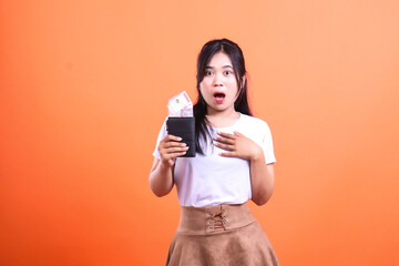 A shocked woman holding a wallet overflowing with Indonesian Rupiah banknotes on orange background