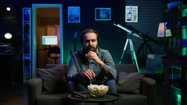 Lazy couch potato person mindlessly watching shows on TV and eating popcorn, paying close attention to screen. Sedentary man devouring snacks while consuming mindless entertainment, camera B