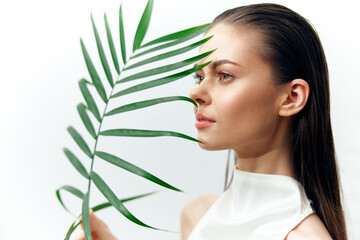 Fototapeta premium Stylish young woman with long hair holding a palm leaf, showcasing minimalistic fashion against a white background.