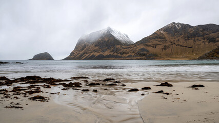 A snow covered mountain stands on Vik Beach under cloudy sky, Lofoten, Norway