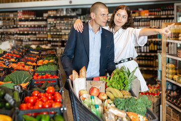 Young male and female customers picking up goods in grocery store, woman pointing at something