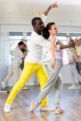 Positive middle-aged woman and Afro-American man engaged in salsa dance in dancehall during workout...