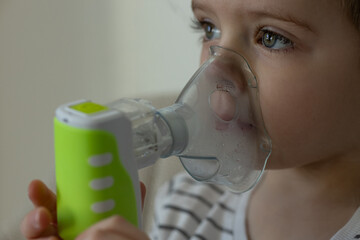 Close-up of a young child with bright eyes using a nebulizer with a green handle for respiratory treatment at home. Pediatric asthma, respiratory therapy and medical equipment for breathing support