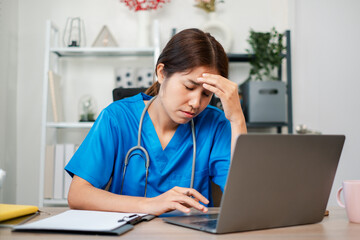 A healthcare worker in blue scrubs looks stressed while working on a laptop in a contemporary office setting.