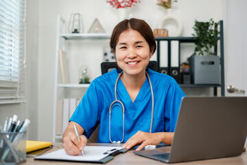 Smiling nurse in blue scrubs working at a desk with a laptop and clipboard in a modern medical office setting.