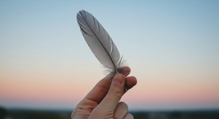 A hand gently holds a delicate feather against a soft sunset.