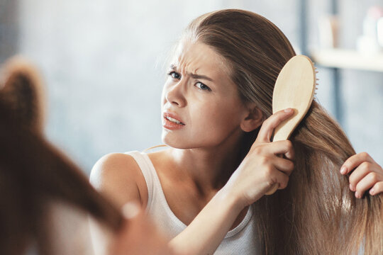 Frustrated young woman trying to brush her tangled hair in front of mirror at bathroom