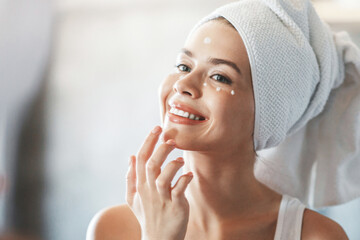 Charming young woman applying under eye cream onto her perfect skin near mirror at home