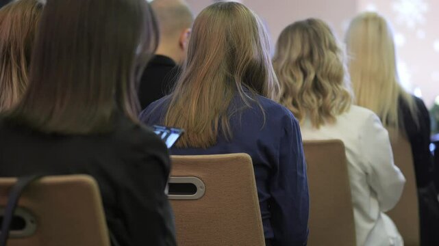Group of people sit in chairs in a room. Scene is one of boredom or disinterest, as the people are not engaged in any activity
