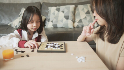Fototapeta premium Asian family, mum, and kid fun playing Go, checkers board game on table in home living room together with love and cheerful, happy weekend relaxing hobby of child and parent, concentrate and compete.