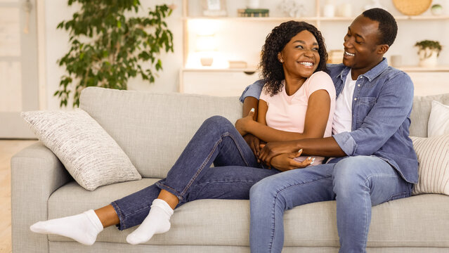 Beautiful african american lovers bonding on couch at living room while spending weekend together at home, copy space. Happy young black man and woman hugging and having pleasant conversation