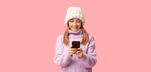 Beautiful mature woman in stylish lilac sweater and knitted hat with mobile phone on pink background