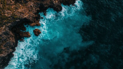 Aerial view of ocean waves crashing against dark rocks. Vivid turquoise and deep blue hues