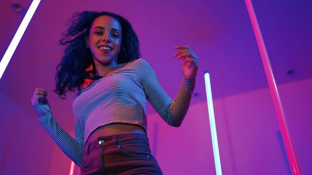 Low angle camera of hispanic dancer looking at camera and dancing to city pop music. Professional attractive hipster moving to hip hop song with purple led or neon light. Stylish cloth. Regalement.