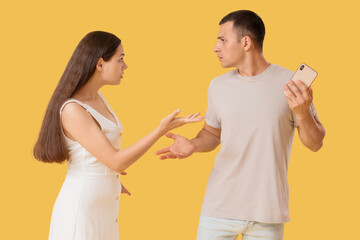 Fototapeta premium Young man with mobile phone and his wife quarreling on yellow background