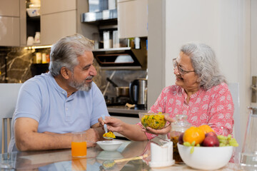 Happy retired senior indian couple eating healthy breakfast together, Elderly husband wife enjoy...