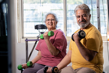 Active indian senior couple doing exercises in gym, Vital elderly people weight lifting training with dumbbells at health club to stay fit.